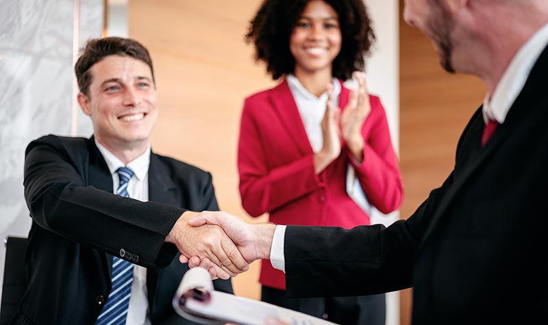 Two professionals in business attire shaking hands over a contract or document while a smiling colleague in a red blazer stands behind them applauding.