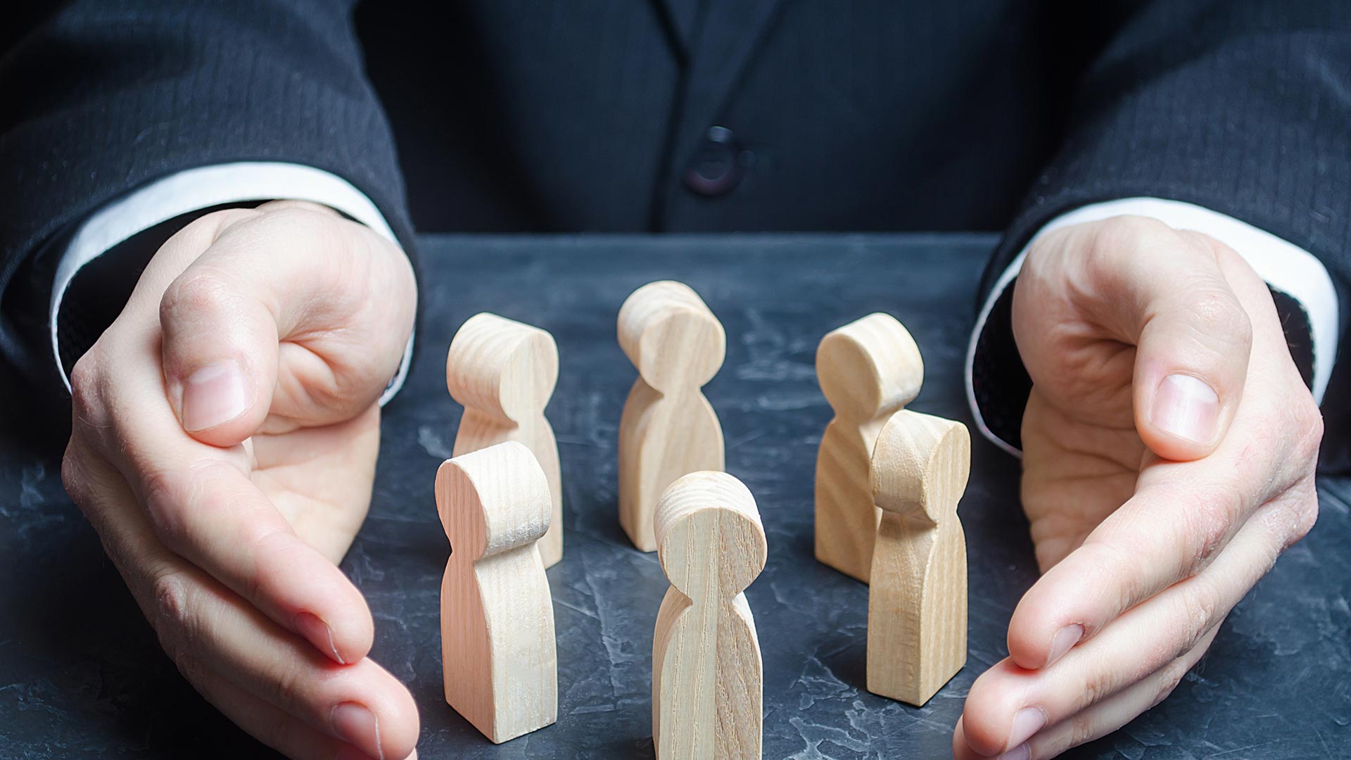 Close-up of a person in a dark suit gently cupping a group of wooden human-shaped figures arranged on a tabletop, symbolizing protection and care.